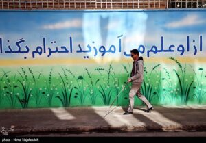 آقای نابینا در حال راه رفتن در حیاط مدرسه است و دیوارنوشته «به کسی که از او علم می‌آموزید احترام بگذارید» در تصویر مشهود است. 